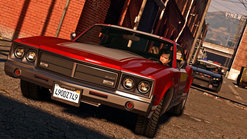 Michael driving a red Declasse Sabre through a sunlit Los Santos alley with a police car behind